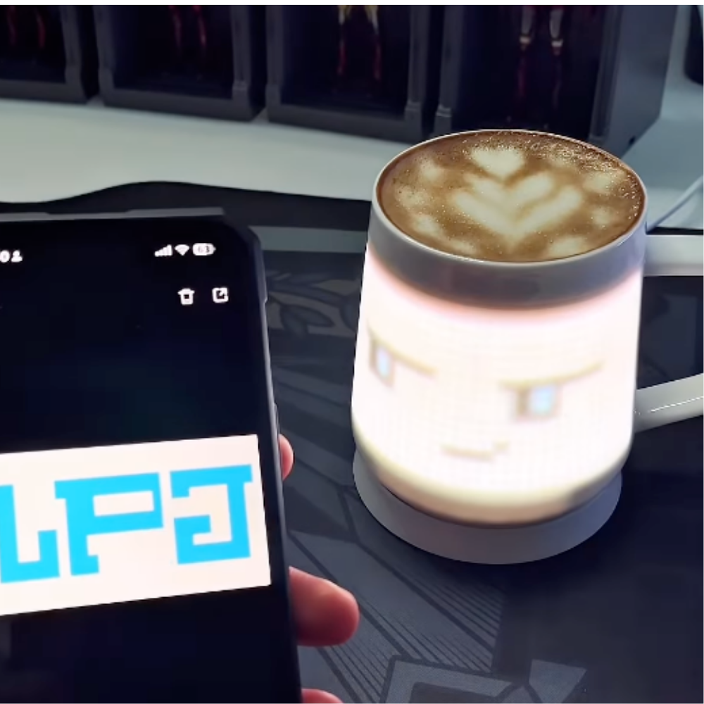 Hand holding a phone with PixelMug app open, syncing pixel display to a ceramic mug with latte art on a gaming desk